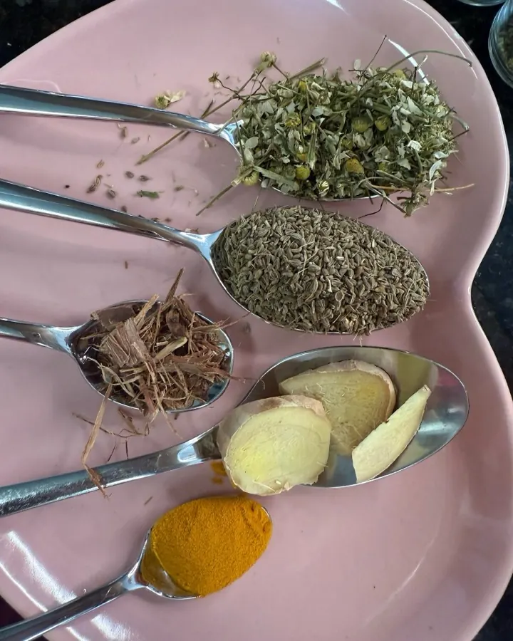 Colheres com ervas secas, raiz de gengibre e cúrcuma sobre um prato rosa